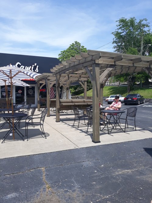 Outdoor dining area with wooden pergola, tables, and a woman sitting at a table, in front of a restaurant.