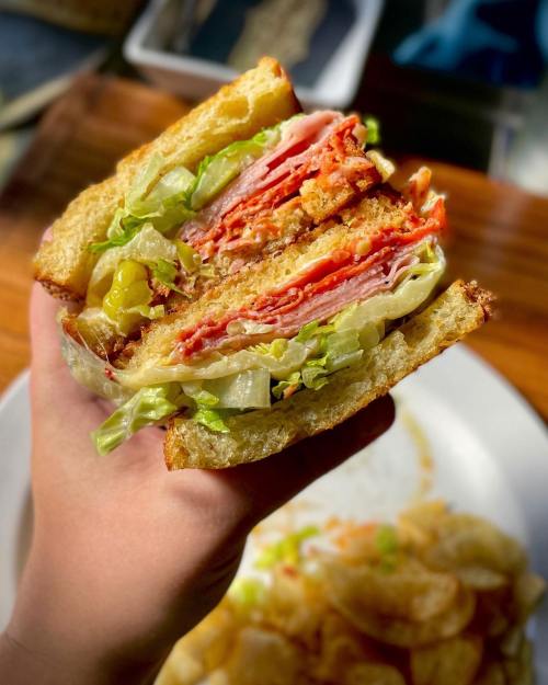 A hand holding a layered sandwich with meats, lettuce, and pickles, alongside a plate of chips.