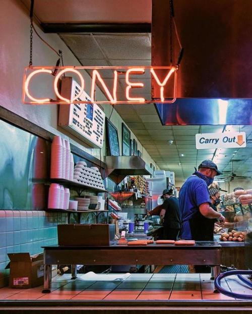 A diner kitchen with a neon "Coney" sign, featuring a cook preparing food and carry-out options visible.