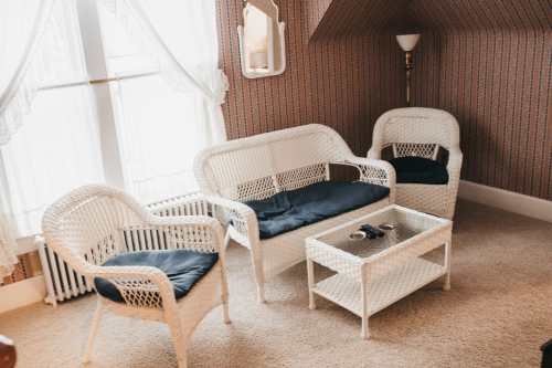 A cozy sitting area with white wicker furniture, including a sofa, two chairs, and a glass coffee table on carpeted flooring.