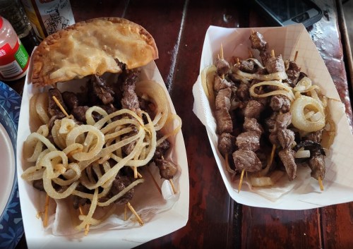 Two trays of skewered meat with grilled onions, one with a pastry on top, served on a wooden table.