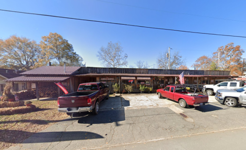 A rustic building with a metal roof, featuring parked trucks and flags, surrounded by trees and clear blue sky.