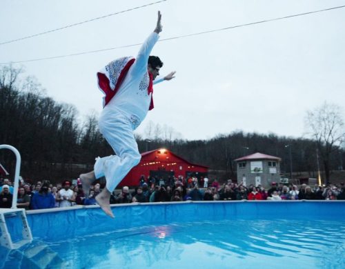 A performer in a white jumpsuit leaps into a pool, with a crowd watching from the edge and buildings in the background.