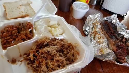 A takeout meal featuring pulled pork, baked beans, mashed potatoes, a slice of bread, and a side of ribs in foil.
