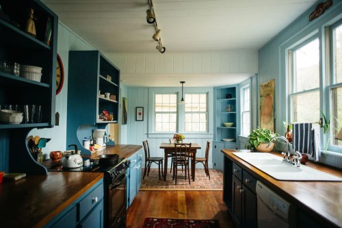Bright kitchen with blue walls, wooden countertops, and a dining area featuring a table and chairs by large windows.