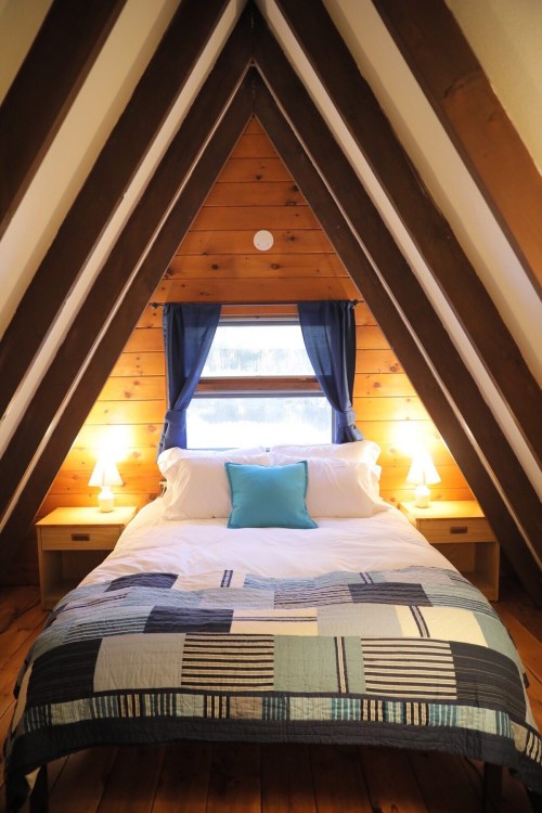 Cozy A-frame bedroom with wooden walls, a double bed, blue accents, and warm lighting from bedside lamps.