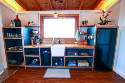 A modern kitchen with wooden shelves, a sink, a fridge, and a window, featuring plants and kitchenware.