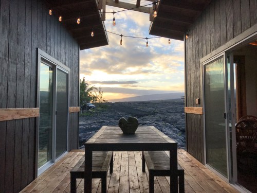 View from a wooden deck between two buildings, featuring a table and a sunset over a rocky landscape.