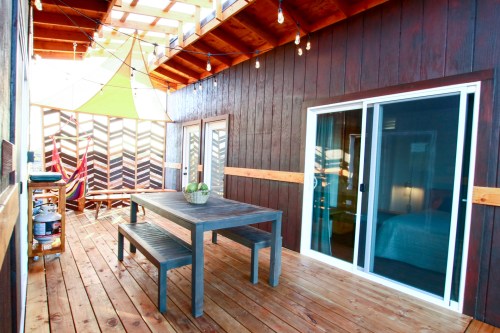 A cozy wooden patio with a table, chairs, and colorful decor, featuring string lights and a hammock.