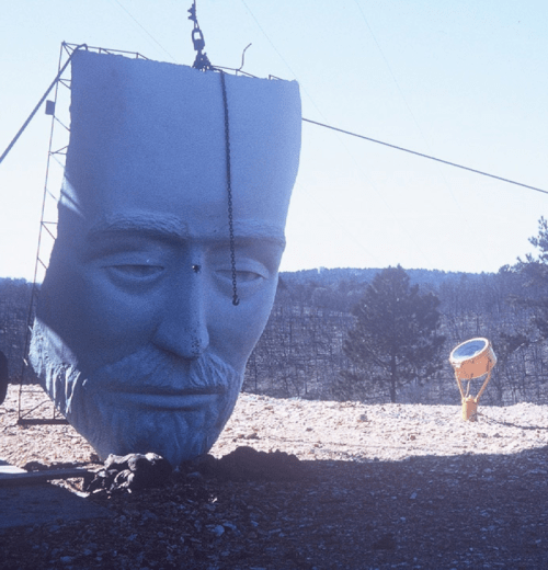 A large, unfinished sculpture of a man's face stands in a barren landscape, with a smaller object nearby.