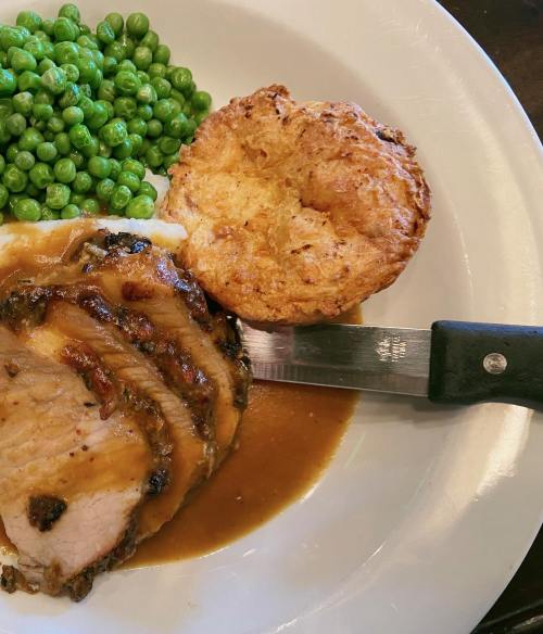 A plate with sliced pork, gravy, green peas, and a biscuit, with a knife resting beside the meat.