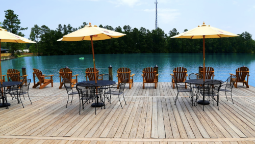 A wooden deck by a turquoise lake, featuring Adirondack chairs and tables under yellow umbrellas.