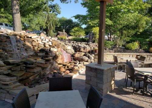 Outdoor seating area with stone waterfall and lush greenery in a sunny setting.