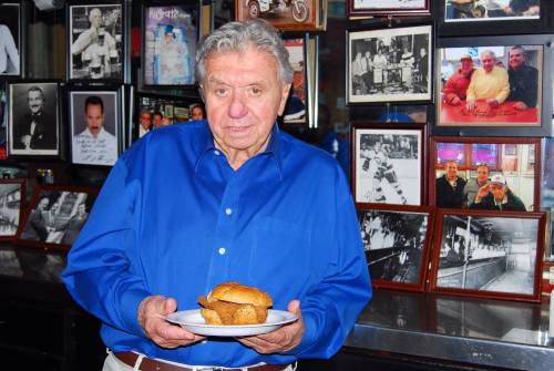 An older man in a blue shirt holds a plate with a burger, standing in front of a wall of framed photos.