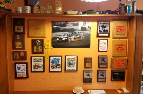 A wooden display featuring various racing trophies, plaques, and a poster of a race car on an orange wall.