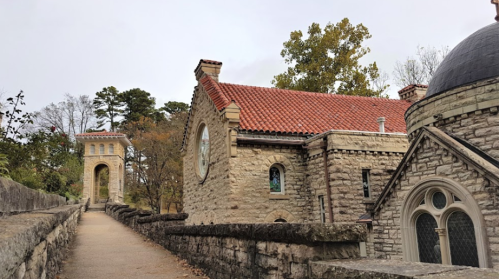 A stone pathway leads to historic buildings with red roofs and an archway, surrounded by trees in a serene setting.