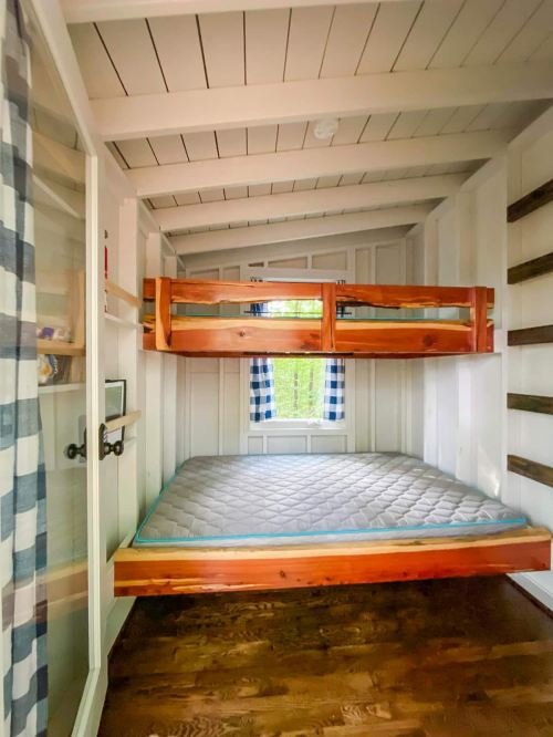 Cozy interior of a small room featuring a wooden bunk bed and a single mattress, with windows letting in natural light.