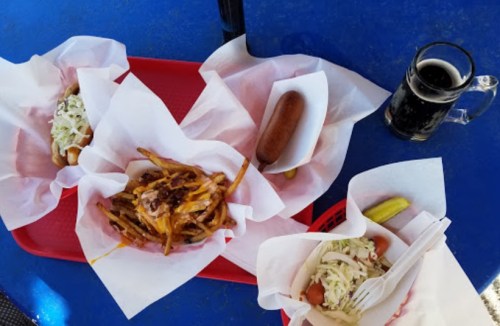 A blue table with three trays of food: hot dogs, fries, and a drink, alongside a pickle.