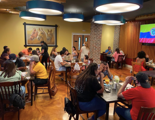 A busy restaurant interior with diners at tables, colorful decor, and a large screen displaying a flag.