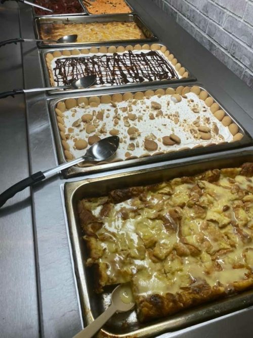 A variety of desserts displayed in trays, including cakes and puddings, with serving spoons on a stainless steel counter.