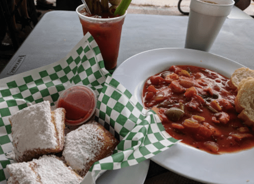 A plate with gumbo, a side of bread, and a basket of powdered sugar beignets on a checkered tablecloth.