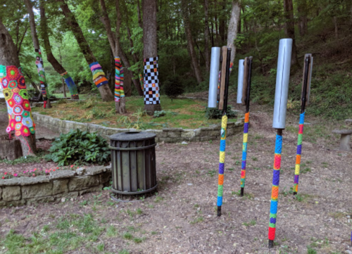 Colorful yarn-wrapped trees and vibrant poles in a wooded park setting, with a trash bin nearby.