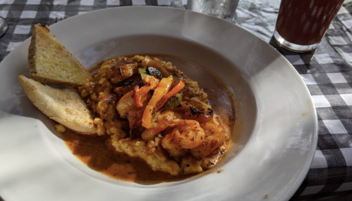 A plate of shrimp and vegetables over creamy grits, served with toasted bread and a drink on a checkered tablecloth.