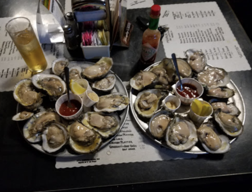 Two plates of fresh oysters with lemon, cocktail sauce, and hot sauce, alongside a glass of beer on a table.