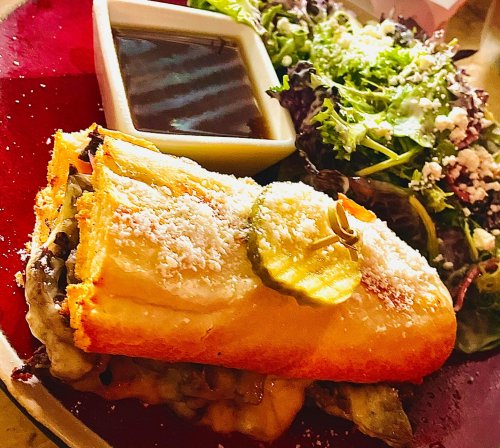 A sandwich topped with pickles and cheese, served with a side salad and dipping sauce on a red plate.