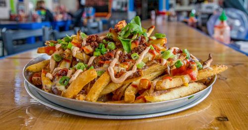 A large platter of loaded fries topped with cheese, bacon, green onions, and drizzled with sauce, served on a wooden table.