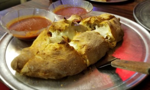 A golden-brown calzone on a metal plate, served with two bowls of red dipping sauce.