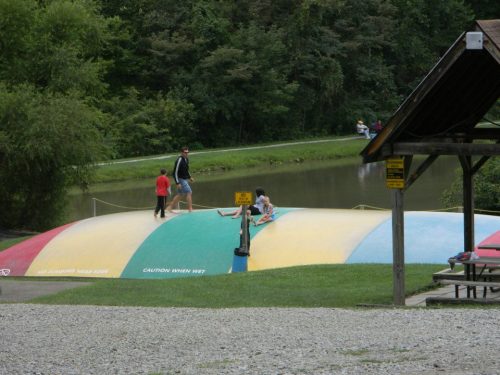 Children and adults play on a colorful inflatable bounce pad near a pond, surrounded by trees and picnic areas.
