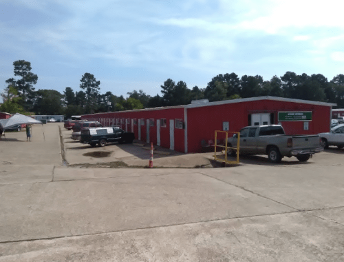 A row of red storage units with parked vehicles and trees in the background under a clear blue sky.
