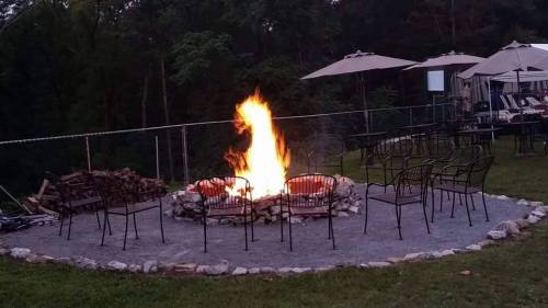 A cozy outdoor fire pit surrounded by chairs, with a tall flame and trees in the background.