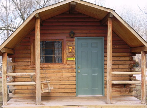 A rustic wooden cabin with a green door, a window, and a porch swing, surrounded by trees.
