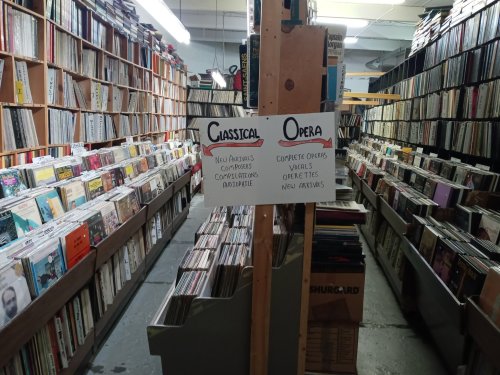A narrow aisle between shelves filled with vinyl records, featuring signs for "Classical" and "Opera" sections.