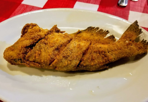 A whole fried fish with crispy skin, served on a white plate against a red and white checkered tablecloth.