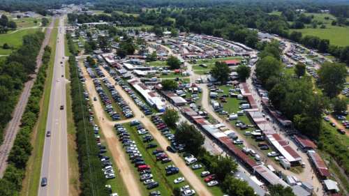 Aerial view of a bustling outdoor market with rows of stalls and parked cars, surrounded by greenery and a road.