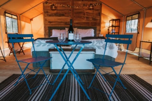 A cozy glamping tent interior with a bed, a small table, two blue chairs, and a bottle of wine with glasses.