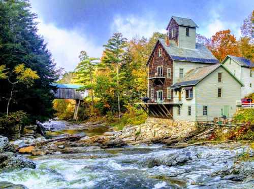 A scenic view of a rustic building by a river, surrounded by autumn trees and a bridge in the background.