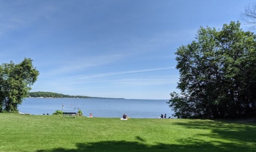 A serene lakeside view with green grass, trees, and people enjoying the water under a clear blue sky.