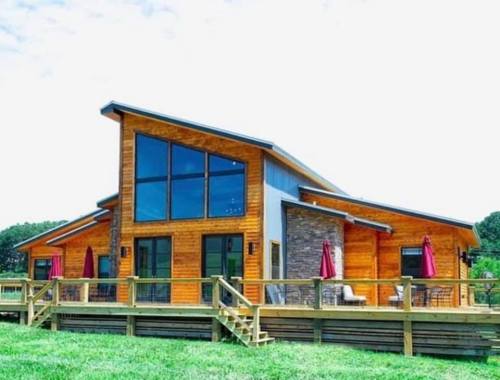 A modern wooden house with large windows, a deck, and red umbrellas, set in a green landscape under a blue sky.
