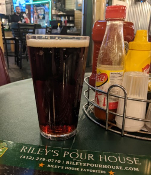 A pint of dark beer sits on a table with condiments and a menu at Riley's Pour House.