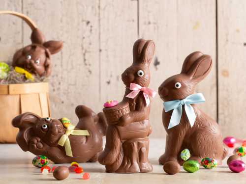 Three chocolate bunnies with colorful bows, surrounded by candy and a basket, set against a rustic wooden background.