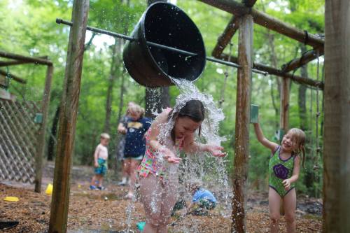 Children playing in a water splash area, with one girl getting drenched from a bucket overhead.