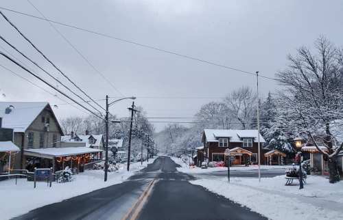 A snowy street scene with quaint houses and festive decorations, surrounded by trees under a gray sky.