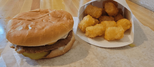 A sandwich with a fried patty and pickles next to a container of crispy tater tots on a wooden surface.