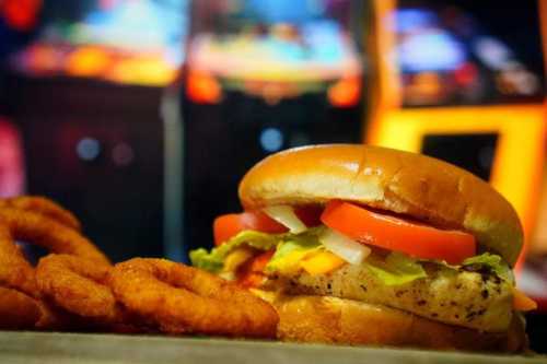 A close-up of a burger with lettuce, tomato, and onion, alongside crispy onion rings, with arcade machines in the background.