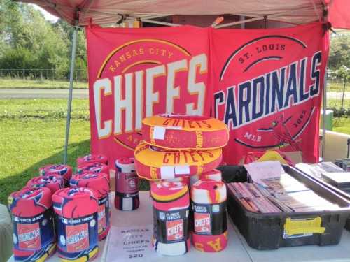 A vendor's booth displays Kansas City Chiefs and St. Louis Cardinals merchandise, including blankets and banners.