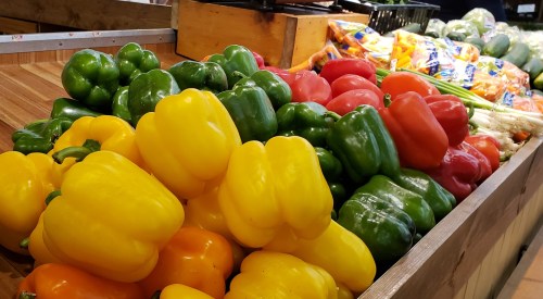 A colorful display of bell peppers in yellow, green, and red, arranged in a wooden crate at a market.